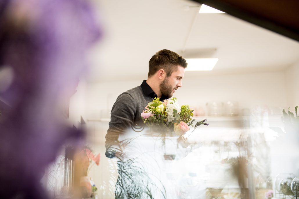Mann bindet Blumenstrauß in heller Floristik-Werkstatt, unscharfe Blüten im Vordergrund.